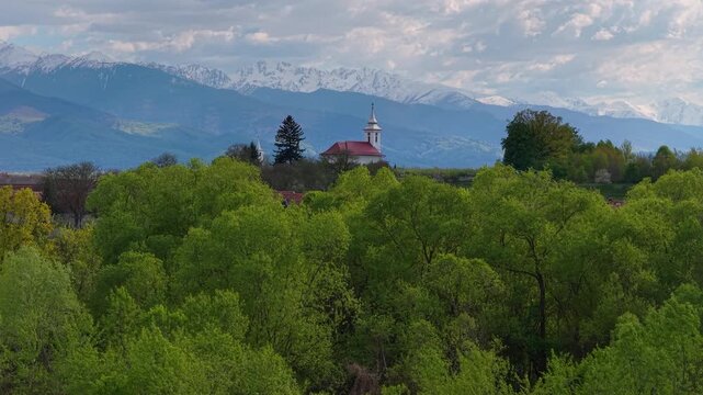 Picturesque white church and swaying green willows in the idyllic rural village of Ucea de Sus against majestic snow capped Fagaras mountains, telephoto low altitude slow forward flight 4K