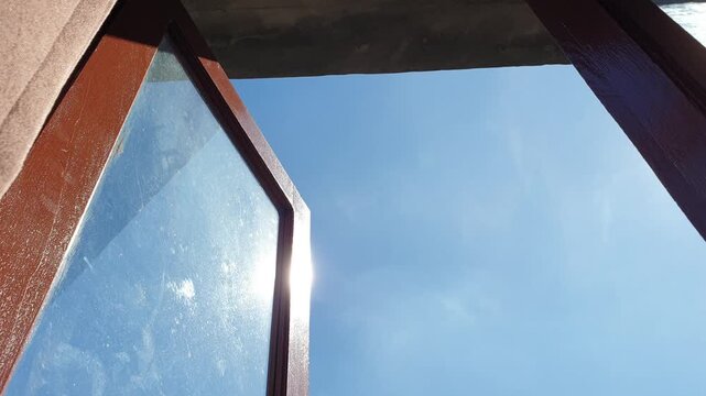 An upward wide shot captures two open wooden window panels framing a gorgeous unblemished blue sky, with brilliant solar flares shining intensely against the dusty left glass surfa
