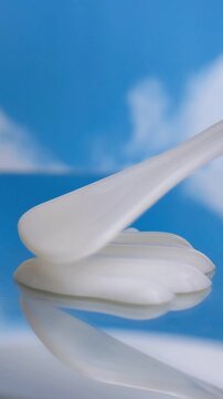Spreading white cream on reflective glass surface using a tool. Blue blurred background. Freshness and UV protection skincare concept.