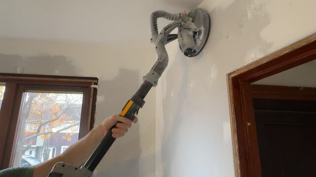 A European man sands a wall with a sander. Wall repair work. Sander in action, medium shot. A construction worker sands drywall and putty. Home renovation and restoration.