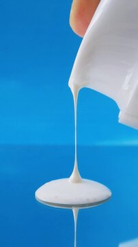 White cream slowly dispensing from bottle on mirror reflection with blue background. Close up view, thick texture for skincare concept.
