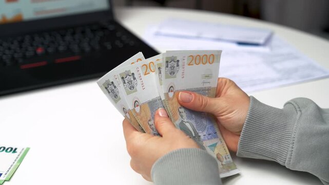 Close-up of woman counting Serbian dinar banknotes while managing household finances on laptop at home