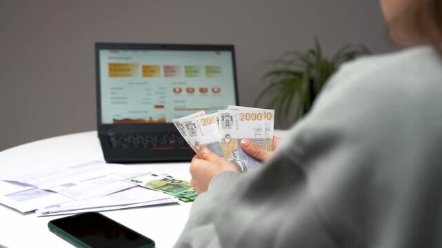 Close-up of woman counting Serbian dinar banknotes while managing household finances on laptop at home