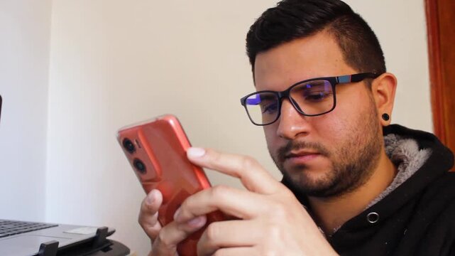 A young Colombian man wearing glasses is using a smartphone to check social media and email. Concept of remote work and technology