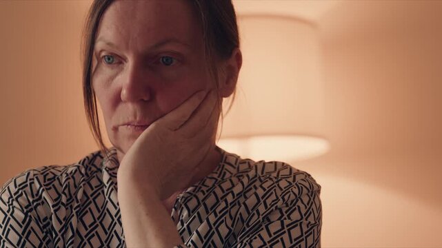 Thoughtful middle-aged woman resting hand against mouth, looking worried in warm indoor light, 4K
