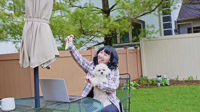 A woman works on her laptop outside in the garden while holding a small dog. She smiles and interacts with the dog, surrounded by green grass and trees.