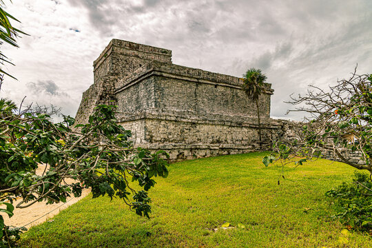 Massive stone El Castillo pyramid on a grassy platform at Chich&eacute;n Itz&aacute;, framed by tropical foliage under cloudy sky