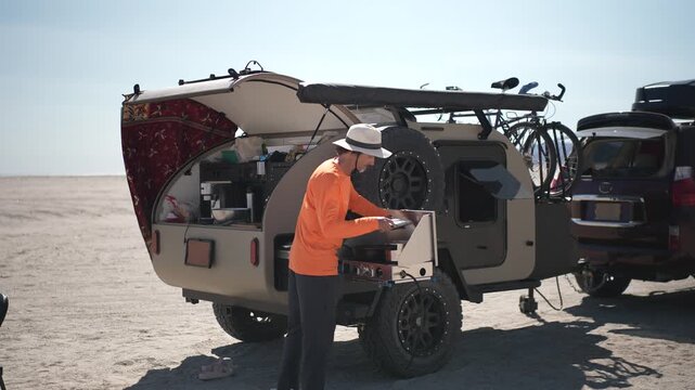 A mature man cooks on a stove outdoors at the beach with a teardrop camper in Baja California Mexico as the sun shines overhead.