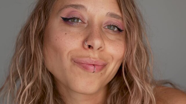 Close-up of young attractive blonde woman with freckles, wavy hair and facial piercings playfully licking her lip. Beauty portrait with flirty expression, pink makeup and soft grey background