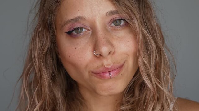 Young attractive blonde woman with wavy hair and facial piercings playfully sticking out her tongue. Close-up beauty portrait with freckles, soft makeup and cheeky confident mood