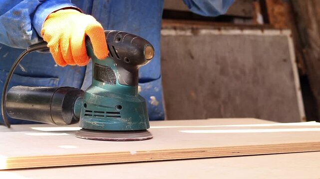 Worker sands wooden surface with random orbital sander. Professional craftsman in protective orange gloves uses orbital sander for finishing plywood. Close-up of woodworking process in workshop