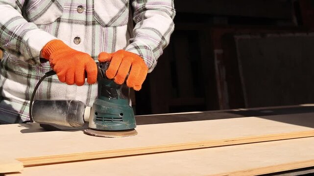 Worker sands wooden surface with random orbital sander. Professional craftsman in protective orange gloves uses orbital sander for finishing plywood. Close-up of woodworking process in workshop