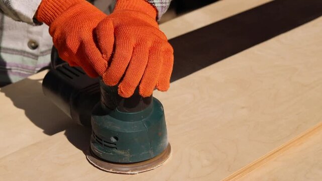 Worker sands wooden surface with random orbital sander. Professional craftsman in protective orange gloves uses orbital sander for finishing plywood. Close-up of woodworking process in workshop