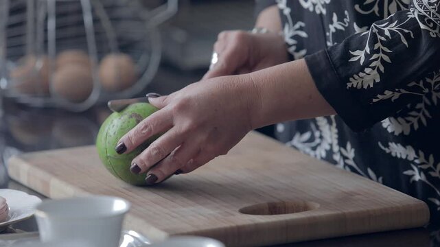 A woman cuts an avocado in half using a knife. In a kitchen, a woman cuts an avocado in half on a cutting board