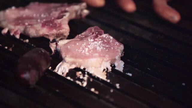 Salting a raw pork chop on a grill. The chop is surrounded by chorizos, other chops, and blood sausage, and it is being seasoned with coarse salt.