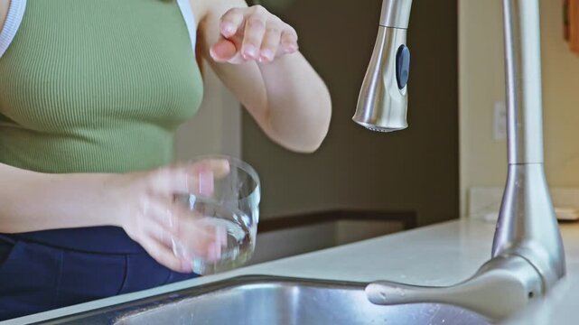 In a kitchen, a person stands at a sink holding a glass with one hand while using the other hand to turn on the faucet. Water flows into the glass as light comes through a nearby window.