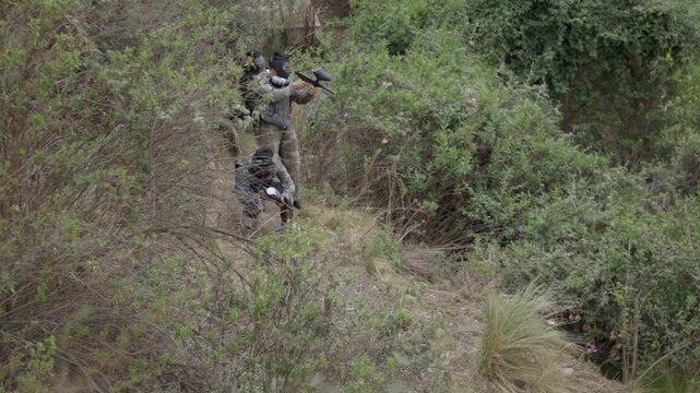 A group of uniformed paintball soldiers armed with paintball guns hide behind some wild bushes, watch the area, and conceal themselves. They are all close together, taking care not to be seen.
