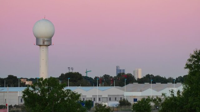 A spherical airport radar dome on a white tower stands against a vivid pink and orange sunset sky, with airport hangars, trees, and a distant city skyline in the foreground