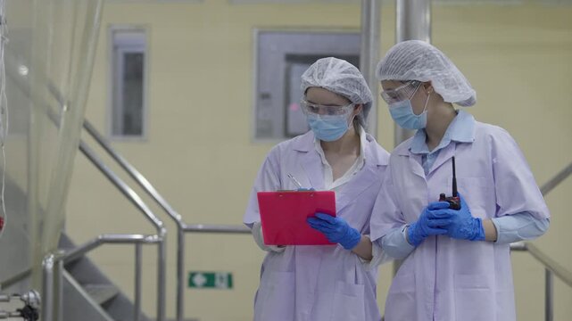 Food factory automated production line, female scientist sterile laboratory inspecting production line with clipboard, safety mask, gloves, hairnet, healthcare worker inspection team sterile corridor