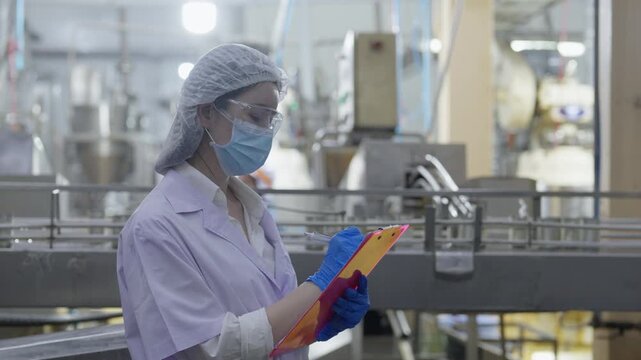 Food factory automated production line, Female factory worker writing checklist in food plant with glove, clipboard, mask, hairnet, and hygiene safety inspection on production line