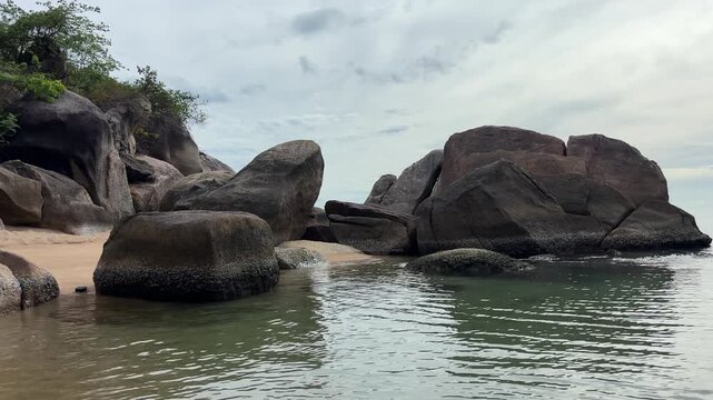 Weathered dark grey rocks rise from turquoise ocean water along the tropical shoreline of Samui Island, Thailand, creating a dramatic coastal landscape under a cloudy sky.