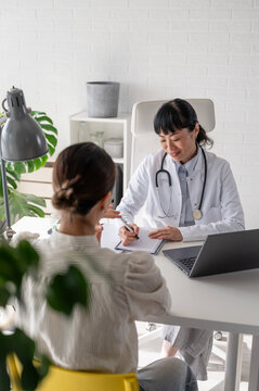 Asian physician writing medical notes during appointment with female patient in bright healthcare office. Preventive care and patient support concept.