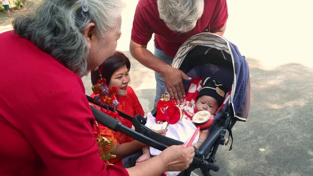 Grandparents took their infant grandson celebrate Chinese New Year visiting Chinese temple and playing with him in a stroller using Chinese doll decorate the stroller and shaking red envelopes as toy.