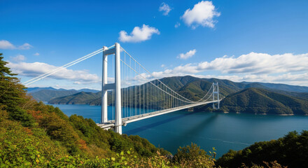 Fototapeta premium Picturesque Suspension Bridge Spanning a Tranquil Bay Surrounded by Lush Green Mountains Under a Bright Blue Sky with Scattered Clouds in Japan
