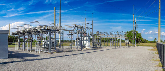 Wide panoramic view of an electrical substation with transformers, high-voltage switchgear, busbars, and utility poles under a blue sky, USA © Andrei Liashchou