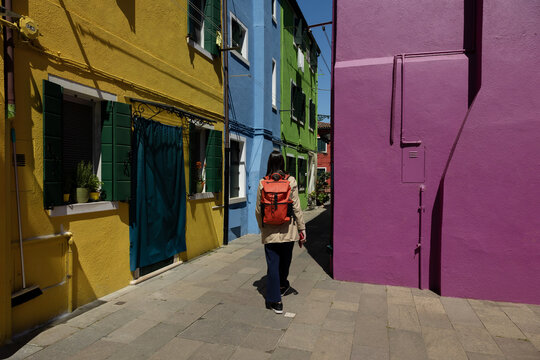 A female tourist with an orange backpack walks among brightly colored houses in Burano, Venice, Italy. The vibrant yellow, blue and purple traditional buildings stand along a sunny paved alleyway.