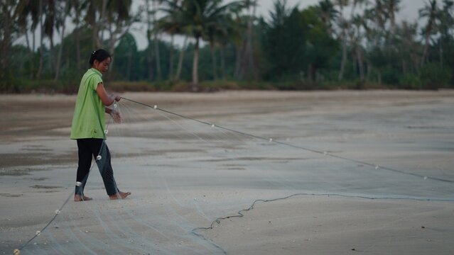 Woman standing barefoot casting a fishing net into the water on a sandy beach