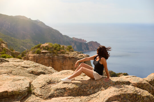 Middle aged woman traveler in a black top and beige shorts relaxing on rocks above the sea at Calanche de Piana, a UNESCO landscape in Corsica. Colors: beige, blue, orange.