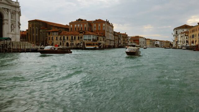Wooden water taxi boats travel on the Grand Canal in Venice, Italy. Historic Venetian architecture and the Basilica di Santa Maria della Salute stand along the water under a cloudy sky