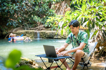 Fototapeta premium Asian man works on a laptop by a tropical waterfall while people swim in the background. Concept of digital nomad, remote work, work-life balance, and business freedom during summer vacation.