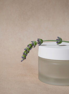 a glass jar of cream with sone lavender flowers on top of it on an empty beige background