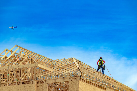 Wide low-angle view of a construction worker standing on a wooden roof truss system with a commercial airplane flying in the blue sky above the building site, USA