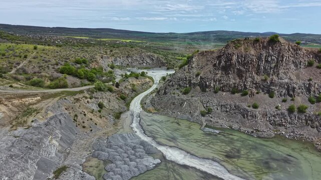 A river flows through a rocky terrain with steep hills surrounding it. Green vegetation appears in certain areas, and the sky shows patches of clouds. The scene captures the natural beauty.
