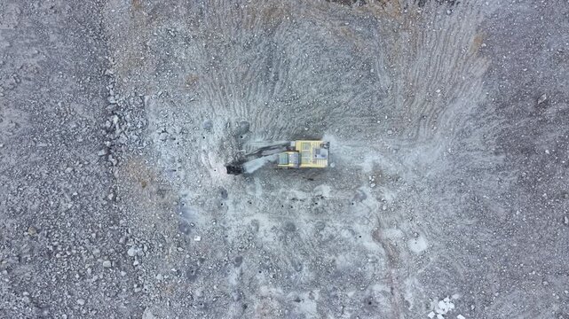 An excavator is digging into a rocky area during the day on a mining site. The earth is uneven and full of stones. The machine is surrounded by gravel and dirt as it works.