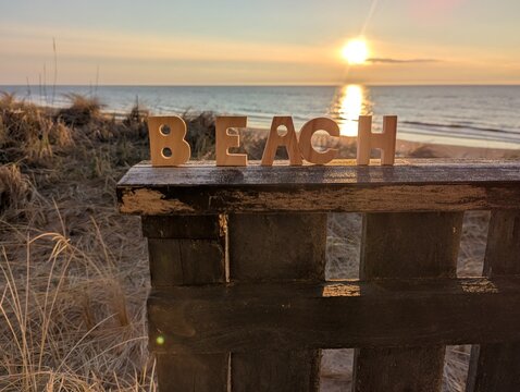 A close-up of small wooden block letters spelling out the word "BEACH," standing on a fence overlooking the sunset over the sea.