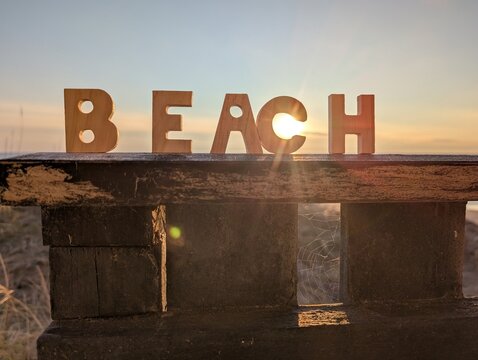 A close-up of small wooden block letters spelling out the word "BEACH," standing on a fence overlooking the sunset over the sea