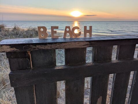 A close-up of small wooden block letters spelling out the word "BEACH," standing on a fence overlooking the sunset over the sea