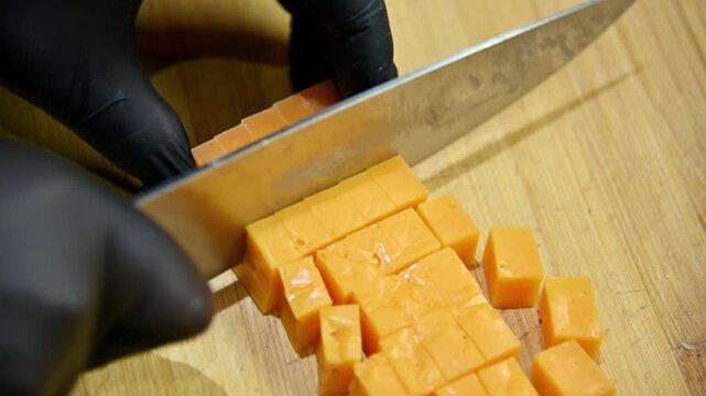 Chef slices cheddar cheese into cubes on wooden cutting board with knife
