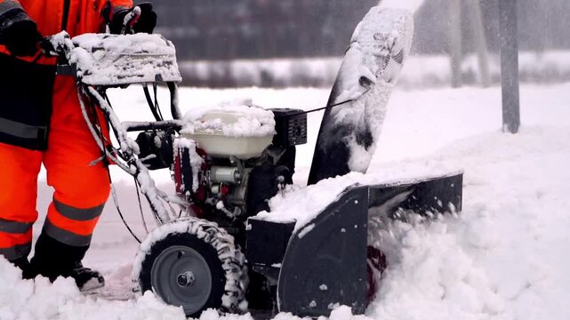 Worker clears snow with heavy machinery. Snow flies as engine hums and blades rotate. Bright orange gear suits stand out in white winter. Scene captures winter maintenance effort