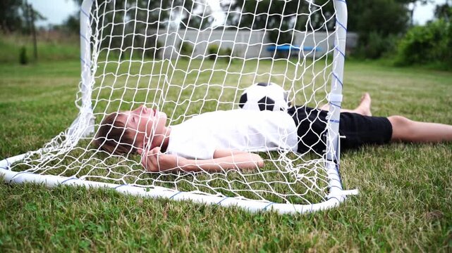 Child rests inside soccer net on grassy field. Child lies peacefully, eyes closed, head on ball. Soft sunlight highlights peaceful moment. Perfect for sports, youth, or family lifestyle scenes
