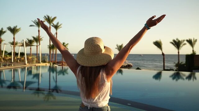 Woman stands poolside, arms raised to sky. Sunset paints warm glow on her back and ocean. Palm trees line tranquil infinity edge. She feels joy, freedom, and peace in the moment