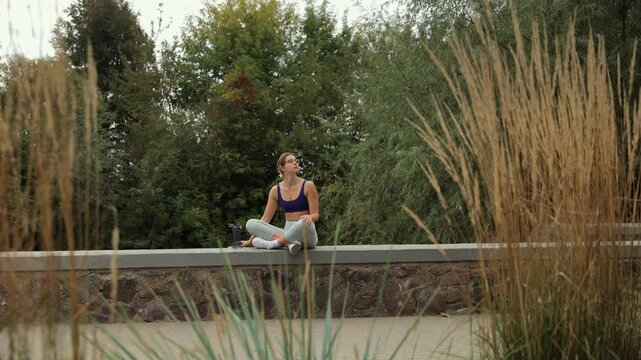 Young athletic woman in sportswear stretching her body while sitting on a stone parapet in a park. She is resting after a workout, checking her phone, and drinking water to stay hydrated
