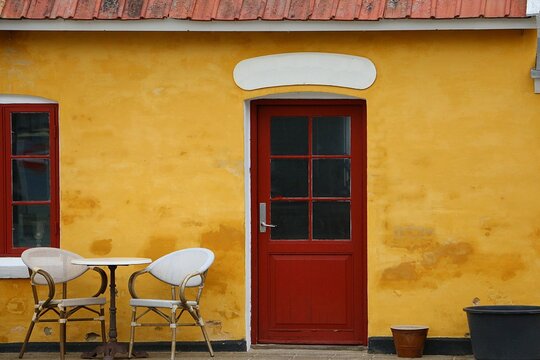 A small, idyllic yellow cottage with a red wooden door and a romantic table and chairs in front of it