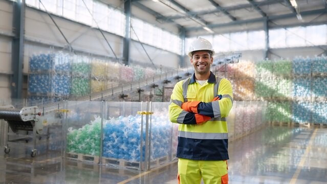 Smiling male worker in recycling plant. Professional man in high visibility uniform and hard hat at waste management facility. Industrial warehouse with plastic bottles background
