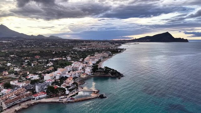 Aerial coastal village at dusk, moody clouds over turquoise sea, row of white houses hugging shoreline,
