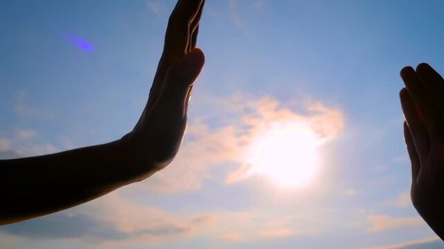 Woman and man silhouettes are giving a high fives hand gesture against the sunset or sunrise sky - close up, sun lens flare. Friendship, celebration, greeting and positive concept